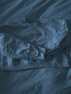 Vit Calvin Klein Jeans hoodie - Säljer en vit hoodie från Calvin Klein Jeans med tryck på bröstet och stor CK-logga i rött och svart på ryggen. Tröjan har ribbade muddar vid ärmslut och nederkant samt en klassisk rund halsringning. Perfekt för dig som gillar stilrena och ikoniska plagg.