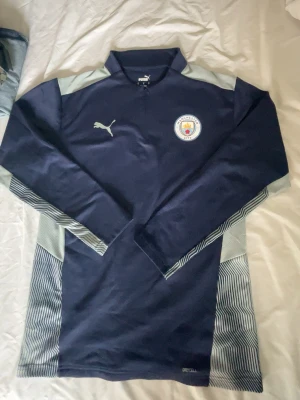Manchester City träningströja Puma - Mörkblå långärmad träningströja från Puma med Manchester Citys klubbmärke på bröstet. Tröjan har ljusgrå detaljer på axlar och ärmar samt randiga partier på sidorna. Tillverkad i funktionsmaterial med DryCell-teknologi.