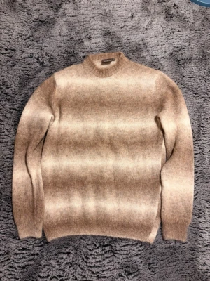 Beige-brun stickad tröja med tonade ränder - Stickad tröja i mjuk ullblandning med breda, tonade ränder i beige och brunt. Tröjan har rund halsringning, långa ärmar och ribbade muddar vid ärmslut och nederkant. Perfekt för lager på lager under kyliga dagar.