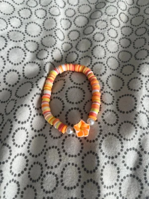Färgglatt pärlarmband med blomma - Armband med platta pärlor i orange, gult och vitt samt två större pärlor i silverfärg. I mitten sitter en orange och gul blomma i polymerlera som ger en söt och unik touch. Perfekt för dig som gillar färg och lekfulla accessoarer.