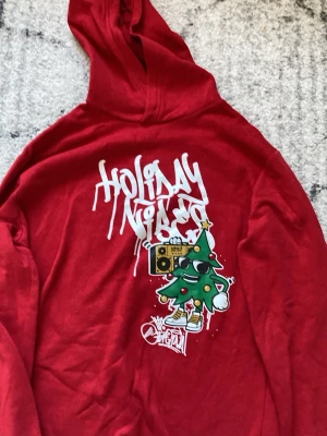 Röd hoodie med julmotiv från H&M - Röd hoodie från H&M med tryck av en cool julgran med solglasögon, sneakers och boombox samt texten 'Holiday Vibes' i graffiti-stil. Hoodien har huva och är gjord i mjukt material, perfekt för dig som vill sticka ut under vintern. Den är aldrig använd bara tvättad. Original pris 280kr