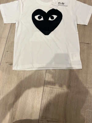 Vit Comme des Garçons Play t-shirt - Säljer en vit t-shirt från Comme des Garçons Play med det ikoniska svarta hjärtat med ögon tryckt på bröstet. Klassisk passform och rund hals. Perfekt statement-plagg för dig som gillar streetwear och vill sticka ut.