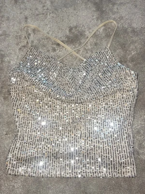 Glittrig paljettopp från NA-KD - Säljer en glittrig topp från NA-KD med tunna axelband och korsad rygg. Beige topp täckt av silverfärgade paljetter som är perfekt på fester eller tjejmiddagar🥰. Materialet är mjukt och stretchigt så passar nog upp till M. Supersnygg till jeans eller kjol! Använd 2 gånger 