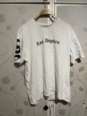 Vit Los Angeles t-shirt Primark - Vit t-shirt från Primark med svart text 'Los Angeles' på bröstet och tryck på ena ärmen. Klassisk rund hals och korta ärmar. Perfekt för en avslappnad streetwear-look.