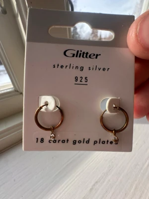 Guldfärgade hoops från Glitter - Snygga hoops i 18 karat guldpläterat sterling silver från Glitter. Örhängena är runda och har en liten pärldetalj som hänger längst ner. Perfekta för dig som gillar stilrena och trendiga smycken.