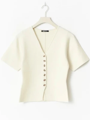 Vit kortärmad kofta från Gina Tricot - Stilren vit kofta från Gina Tricot med korta ärmar och djup V-ringning. Koftan har dekorativa knappar framtill i sköldpaddsmönster och är tillverkad i ett mjukt, stickat material. Perfekt att matcha med jeans eller kjol för en trendig look.