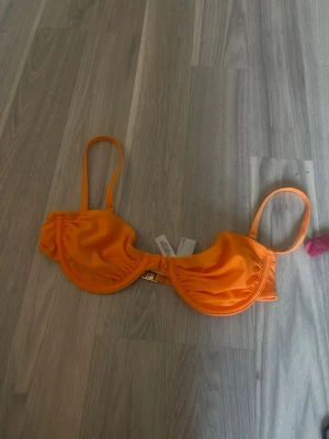 Orange bikiniöverdel med bygel - Snygg orange bikiniöverdel med bygel och tunna axelband. Modellen har en metallspänne framtill och ger ett trendigt och fräscht intryck. Perfekt för soliga dagar på stranden eller vid poolen.