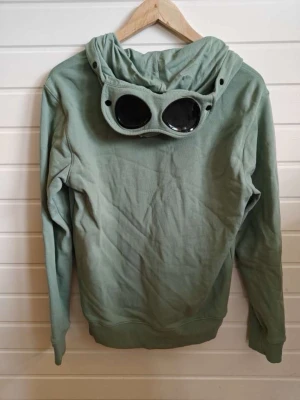 Grön hoodie från C.P. Company - Säljer en grön hoodie från C.P. Company med dragkedja framtill och två fickor. Huvan har de ikoniska glasögon-linserna som ger en unik look. Tillverkad i mjukt bomullsmaterial och har ribbade muddar vid ärmslut och nederkant. Perfekt för dig som vill sticka ut. En knapp som man spänner luvan med har lossnat men det är bara att sy fast den igen😁