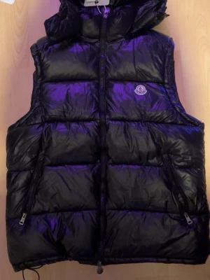 Svart dunväst från Moncler - Snygg svart dunväst från Moncler med glansig finish och ikonisk logga på bröstet. Västen har dragkedja framtill, två fickor med dragkedja och en avtagbar huva. Insidan har ett coolt serietidningsmönster. Perfekt för lager på lager och streetstyle. Den kan också passa S