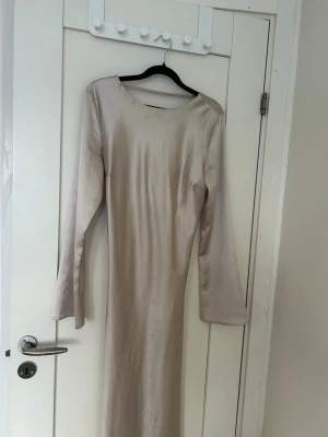 Beige långklänning med knyt fram - Stilren beige långklänning i satinliknande material med lång ärm och snygg v-ringning. Klänningen har ett dekorativt knyt framtill som ger en feminin touch. Perfekt för dig som gillar minimalistisk och elegant stil.