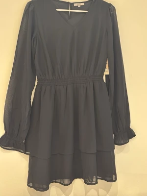 Svart långärmad kortklänning - Svart långarmad kortklänning med volanger. Helt oanvänd och lappen är kvar. I storlek 158/164