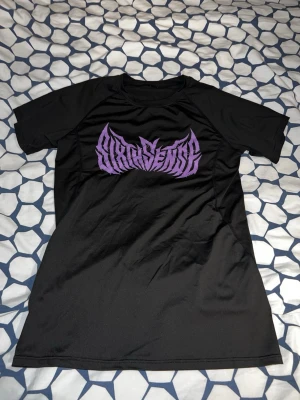 Svart t-shirt med lila tryck - Snygg svart t-shirt med ett stort grafiskt tryck i lila på bröstet. T-shirten har korta ärmar och en klassisk rund hals. Materialet känns som polyester vilket ger en sportig vibe och passar perfekt till en avslappnad stil.