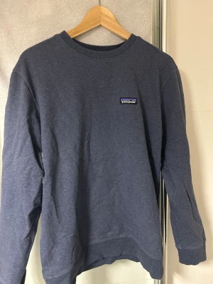 Mörkblå/Grå sweatshirt från Patagonia - Mörkblå sweatshirt från Patagonia med klassisk rund halsringning och diskret logga på bröstet. Tröjan har en avslappnad passform och är tillverkad i mjukt bomullsmaterial. Perfekt för en chill och stilren look. Köpt på herravdelningen men passar unisex. Ordinarie pris 1000kr