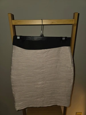 Beige kortkjol med struktur - Säljer en beige kortkjol med svart midjeresår och vågig struktur i tyget. Kjolen har en enkel, rak passform och är perfekt för dig som gillar stilrena plagg med en twist. Materialet känns mjukt och stretchigt, vilket gör den bekväm att bära.