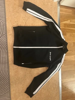 Palm Angels svart track jacket - Säljer en svart track jacket från Palm Angels med vita ränder längs ärmarna och vit dragkedja. Jackan har ståkrage, två fickor med dragkedja och Palm Angels-logga på bröstet. Perfekt för en sportig och trendig look. Ordinarie pris 3000kr, men säljer för 1000kr (för liten för mig). Vid seriösa köp kontakta mig för mer bilder och frågor!