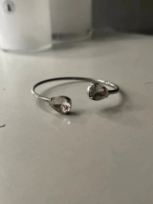 Silverfärgat armband med droppformade stenar - Snyggt armband i silverfärgad metall med två stora, klara droppformade stenar i varje ände. Fint skick och pris kan diskuteras 💞