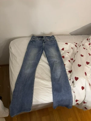 Blå bootcut jeans från True Religion - Säljer ett par blå bootcut jeans från True Religion med klassiska kontrastsömmar och broderade bakfickor. Jeansen har en snygg tvätt och är tillverkade i denim. Perfekta för dig som gillar en avslappnad men trendig stil.