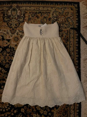 Vit broderad kjol från Zara - Söt vit kjol från Zara med bred smock i midjan och broderade hålmönster över hela tyget. Kjolen har en lätt utställd form och avslutas med vågig kant nertill. Perfekt för en fräsch och somrig look.
