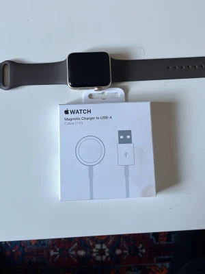 Apple Watch med grått silikonarmband - Snygg Apple Watch med rektangulär svart skärm och grått silikonarmband. Klockan har en elegant metallram och kommer med en magnetisk laddare till USB-A. Perfekt för dig som vill ha koll på notiser, träning och hälsa direkt på handleden. Airpodsen gen 2 (nya direkt från lådan) går att köpa till för 800kr