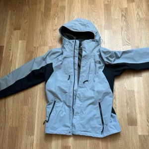 Grå vindjacka från The North Face - Snygg grå vindjacka från The North Face med svarta detaljer. Jackan har huva med dragsko, flera dragkedjeförsedda fickor och justerbara ärmslut. Perfekt för blåsiga dagar och outdoor-äventyr. Materialet är slitstarkt och vattenavvisande.