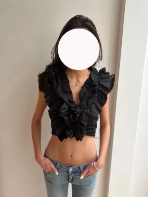 Svart croppad volangblus🖤 - Säljer en svart croppad blus med stora volangdetaljer framtill och baktill. Toppen är ärmlös och har en djup v-ringning. Materialet känns som polyester Så snygggg!! 🖤🥰