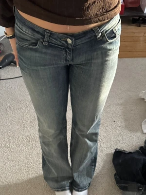 Super low waist bootcut jeans Nelly - Säljer ett par blå bootcut jeans med låg midja och klassisk femficksmodell. Jeansen har slitningar och tvättade detaljer för en vintagekänsla. Snygg broderad detalj på bakfickan. Perfekta till en avslappnad och trendig look.