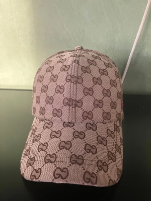 Beige keps med GG-mönster från Gucci - Säljer en beige keps från Gucci med det klassiska GG-monogrammet i brunt över hela kepsen. På sidan finns en grön och röd rand som signaturdetalj. Kepsen har böjd skärm och justerbar rem bak. Materialet är canvasliknande tyg.