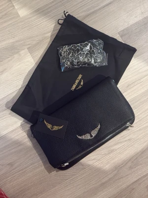 Svart kuvertväska Zadig & Voltaire - Snygg svart kuvertväska från Zadig & Voltaire i strukturerat läder med silverfärgad vinge framtill. Väskan har dragkedja och medföljer gör en svart dustbag samt en kedja i silverfärg som kan användas som axelrem. Perfekt för dig som gillar edgy detaljer.