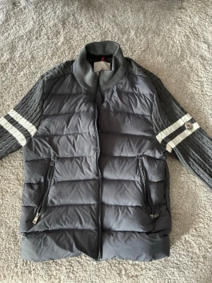 Grå Moncler dunjacka med stickade ärmar - Snygg grå dunjacka från Moncler med stickade ärmar i mörkgrått och vita ränder. Jackan har två dragkedjefickor framtill och ribbad krage. Moncler-logga på ena ärmen. Perfekt för kalla vinterdagar och riktigt stilren look.