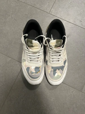 Valentino camo sneakers i pastell - Snygga sneakers från Valentino med pastellfärgat camo-mönster i beige, grått och grönt. Skorna har vita läderdetaljer, vita snören och svarta innersulor. Sidan har nitar vid hälen och sulan är beige. Perfekta för dig som vill sticka ut med stil.