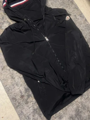 Svart windbreaker Moncler - Snygg svart vindjacka från Moncler med huva och dragkedja framtill. Jackan har Moncler-logga på bröstet och detaljer i rött, vitt och blått på insidan av huvan. Perfekt för blåsiga dagar och enkel att matcha.Perfekt nu när våren börja närma sig.Pris kan diskuteras!!
