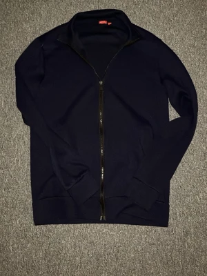 Mörkblå ziptröja från RYVLS - Mörkblå ziptröja från RYVLS med hel dragkedja framtill och ståkrage. Tröjan har ribbade muddar vid ärmslut och nederkant, perfekt för en stilren och enkel look. Materialet känns mjukt och bekvämt mot huden. Storlek 158-164 och passar TTS, har du några frågor är det bara att skriva💫
