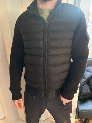 Svart dunväst från Moncler - Snygg svart dunväst från Moncler med quiltad front och stickade ärmar. Västen har hög krage, dragkedja framtill och en ficka med Moncler-logga på ärmen. Perfekt för lager-på-lager och ger en clean streetwear-look.