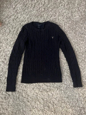 Mörkblå kabelstickad tröja GANT - Snygg mörkblå kabelstickad tröja från GANT med rund halsringning och diskret logga broderad på bröstet. Tröjan har långa ärmar och ribbade muddar vid ärmslut och nederkant. Perfekt för en klassisk och stilren look.