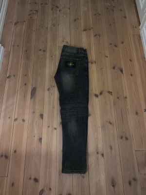 Svarta jeans från Stone Island - Säljer ett par svarta jeans från Stone Island med klassisk patch på bakfickan. Jeansen har en slim fit passform och snygga slitningar framtill för en trendig look. Tillverkade i denim med fem fickor och knappgylf.