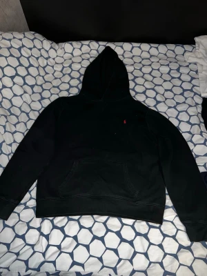 Svart hoodie från Polo Ralph Lauren - Klassisk svart hoodie från Polo Ralph Lauren med röd broderad logga på bröstet. Hoodien har stor huva, magficka och ribbade muddar vid ärmslut och nederkant. Tillverkad i mjukt bomullsmaterial som är perfekt för chill dagar. Storlek xl i barnstorlek så m vanligt 
