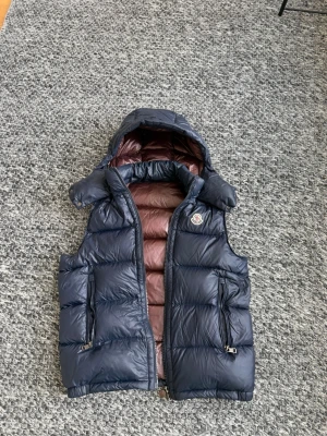Mörkblå dunväst från Moncler - Snygg mörkblå dunväst från Moncler med avtagbar huva och brunt innerfoder. Västen har två dragkedjefickor framtill och klassisk Moncler-logga på bröstet. Perfekt för lager-på-lager och håller dig varm under kyliga dagar.