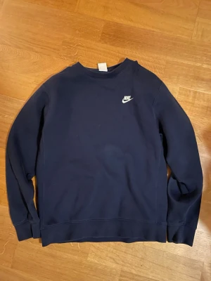 Mörkblå sweatshirt från Nike - Klassisk mörkblå sweatshirt från Nike med rund halsringning och vit Nike-logga på bröstet. Tröjan har långa ärmar och ribbade muddar vid ärmslut och nederkant. Perfekt för en avslappnad och sportig stil.