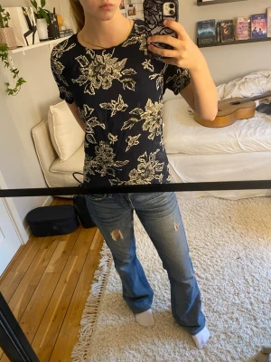 Mörkblå blommig blus - Snygg mörkblå blus med stort blommigt mönster i beige. Klassisk rund halsringning och korta ärmar. Skön passform och lätt att matcha med jeans eller kjol. Är relativt gammal men ändå bra skick.