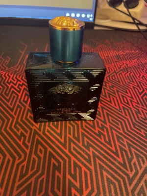 Versace Eau de Toilette blå flaska - Snygg parfym från Versace i en mörkblå, fyrkantig glasflaska med elegant mönster och guldblått lock. Flaskan har Versaces ikoniska Medusa-detalj på framsidan och känns riktigt lyxig och stilren. Perfekt för dig som gillar exklusiva dofter och snygg design. Den har ungefär 23 ml kvar