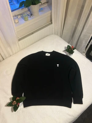 Svart sweatshirt från Ami Paris - Säljer en svart sweatshirt från Ami Paris med deras ikoniska lilla vita hjärtemblem broderat på bröstet. Tröjan har rund halsringning, långa ärmar och ribbade muddar. Perfekt för en clean och stilren look.