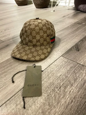 Beige keps från Gucci med GG-mönster - Säljer en beige keps från Gucci med det klassiska GG-monogrammönstret över hela kepsen. På sidan finns en grön och röd rand som signaturdetalj. Kepsen har böjd skärm och justerbar rem bak. Perfekt för dig som vill ha en lyxig och trendig accessoar. Skicka för fler bilder!!