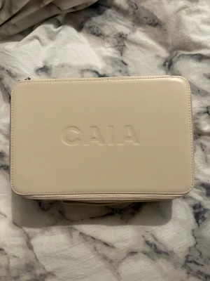 Beige necessär från CAIA - Snygg beige necessär från CAIA i rektangulär form med dragkedja runt om. Tillverkad i ett slätt material med CAIA-loggan präglad på locket. Invändigt finns ett fack i locket för extra förvaring. Perfekt för smink eller hudvårdsprodukter. Helt ny!!!