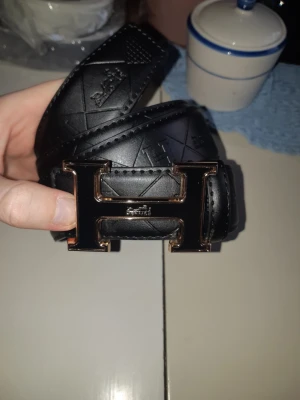 Svart skärp med H-spänne - Snyggt svart skärp från Hermès med stort H-format metallspänne. Skärpet har diskreta mönster i lädret och känns riktigt lyxigt. Perfekt accessoar för att lyfta vilken outfit som helst.