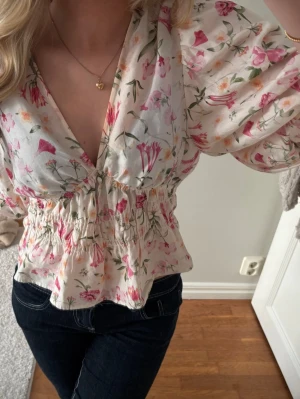 Blommig blus med puffärmar - Söt blommig blus i vitt med rosa, gröna och orange detaljer. Djup v-ringning, puffiga ärmar och smockad midja. Perfekt för vår och sommar, matcha med jeans eller kjol.