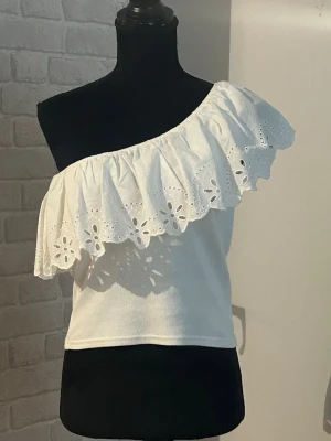 Vit oneshoulder topp med volang - Snygg vit oneshoulder topp med bred volangkant i broderad spets och hålmönster. Jag har använt den ganska mycket och är därför med på att sälja den ännu billigare, men den är i jätte fint skick utan några fläckar eller hål.