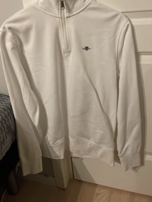 Vit halfzip hoodie från Gant - Snygg vit hoodie från Gant med halv dragkedja och liten broderad logga på bröstet. Tillverkad i mjukt material och har ribbade muddar vid ärmslut och nederkant. Perfekt för en clean och stilren look.