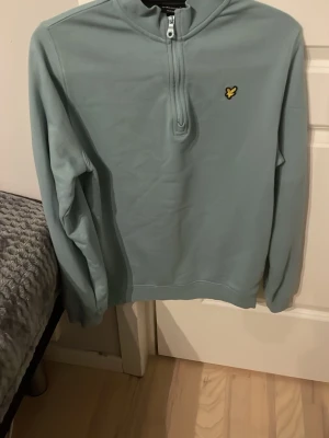Ljusblå halfzip hoodie från Lyle & Scott - Snygg ljusblå hoodie från Lyle & Scott med halv dragkedja och deras klassiska gula logga på bröstet. Hoodien har en enkel och clean design utan fickor och är tillverkad i mjukt material som känns skönt mot huden. Perfekt för en avslappnad och stilren look.