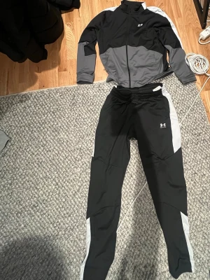 Under Armour svart träningsset - Snyggt träningsset från Under Armour med svart och grå zip-jacka och matchande byxor. Setet har vita detaljer längs ärmar och ben, samt dragkedja framtill på jackan. Perfekt för gymmet eller löprundan. Materialet är troligtvis polyester för bra andningsförmåga.