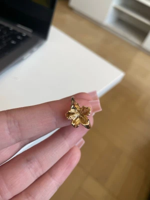 Guldfärgad ring med fyrklöver - Säljer en snygg guldfärgad ring med en stor fyrklöver som detalj. Ringen har en blank finish och klövern är i en cool, lite skulptural design. Perfekt för dig som gillar unika accessoarer och vill sticka ut lite extra. Fick den i present men använder tyvärr inte guld, var svårt att sätta pris då jag inte vet vart den är ifrån men den verkar vara i bra skick.